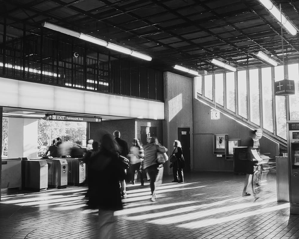 Black-and-white photograph of a BART transit station, with commuters in motion blur passing through turnstiles under the morning light